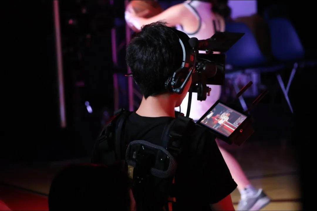 Seen from behind, a camera operator wearing a headset and a body rig films a school event, with a small monitor mounted beside the camera.