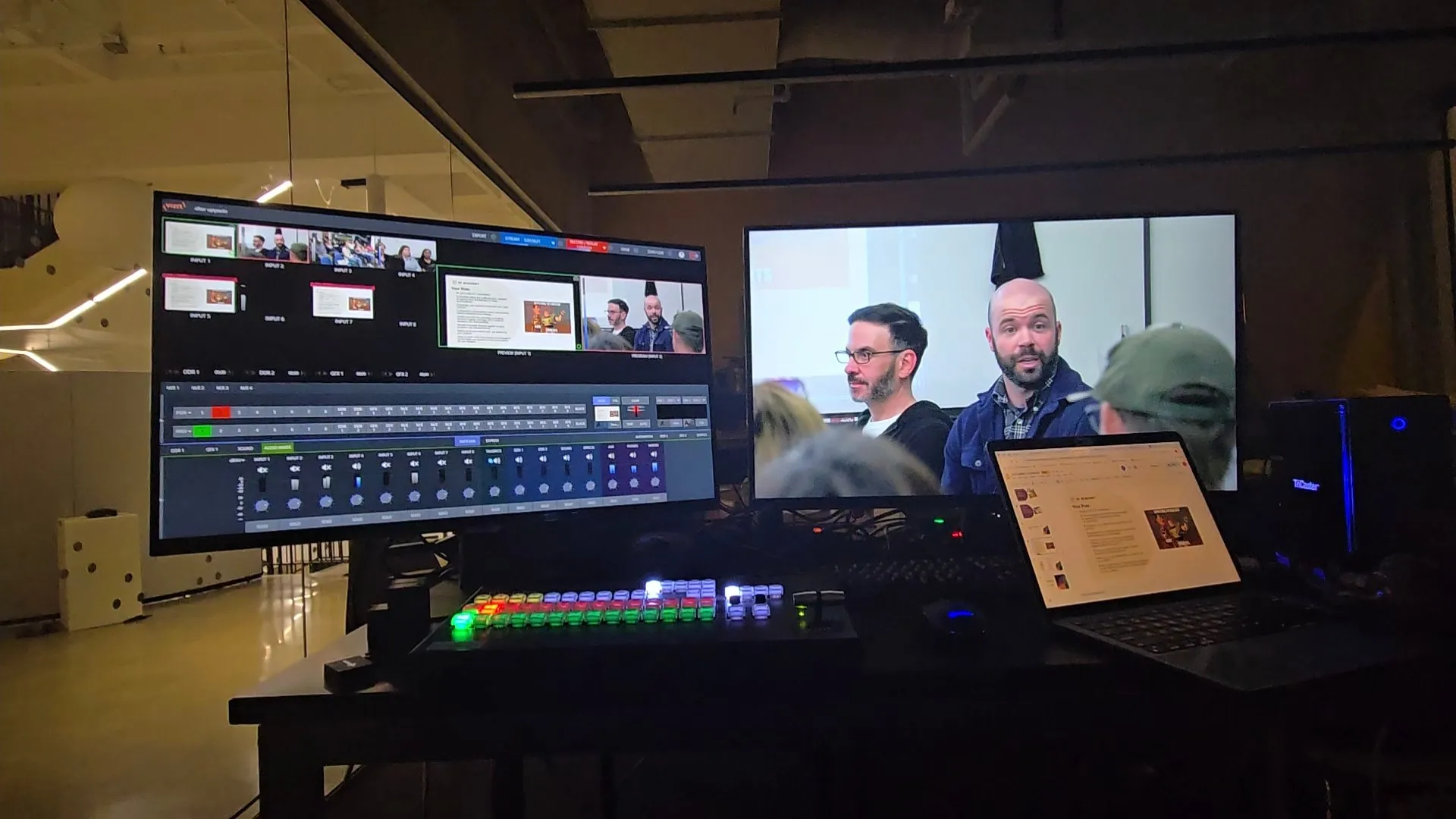 A livestream control setup in a dark room, with a video switcher with lit buttons in front of a monitor showing the camera inputs, a second screen showing two speakers, a laptop with slides and a TriCaster unit.