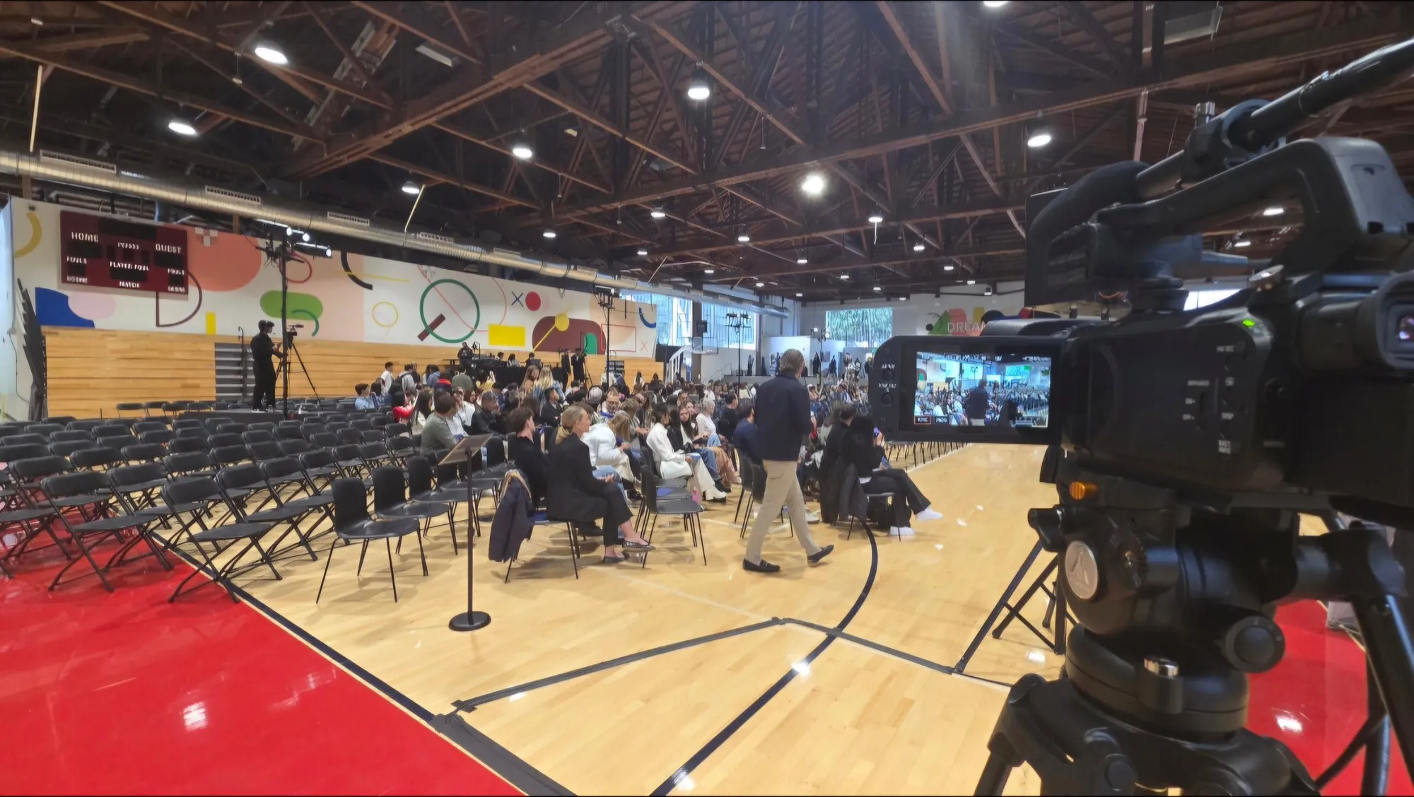 A camera on a tripod in the foreground of the school gym, facing rows of folding chairs and a seated audience, with another camera operator on the far side of the room.