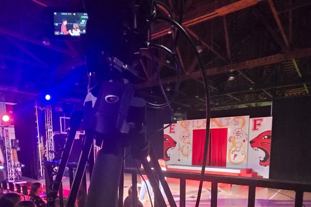 A camera on a tripod, with cables running down it, looks out toward a stage with a Wildcats backdrop. Its small screen shows two performers.